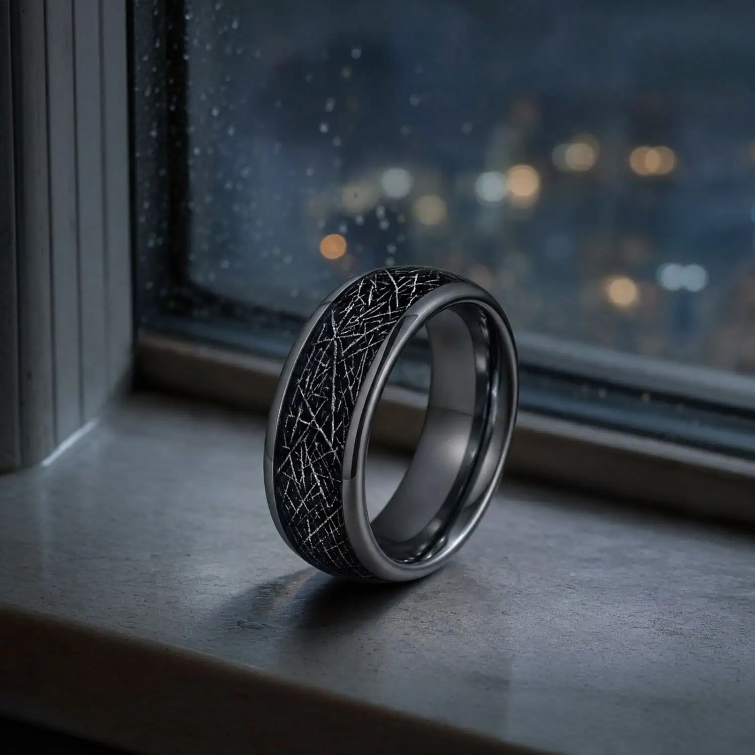 Silver tungsten ring with a black meteorite-style inlay standing on a windowsill beside rain-speckled glass, with blurred city lights at night.