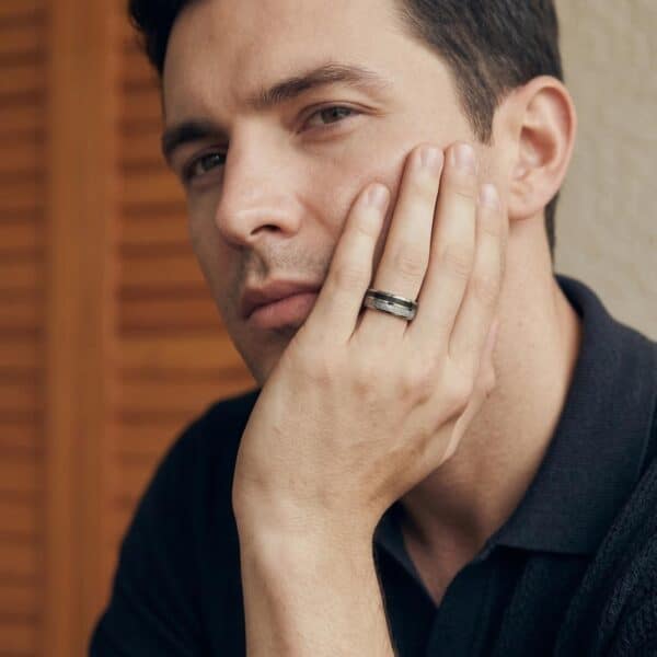 In a thoughtful pose, a man sits with his hand held up to his face. Looking out to one side of the camera, the man is wearing a silver ring. The ring is made with tungsten and features faux meteorite and black carbon fibre.