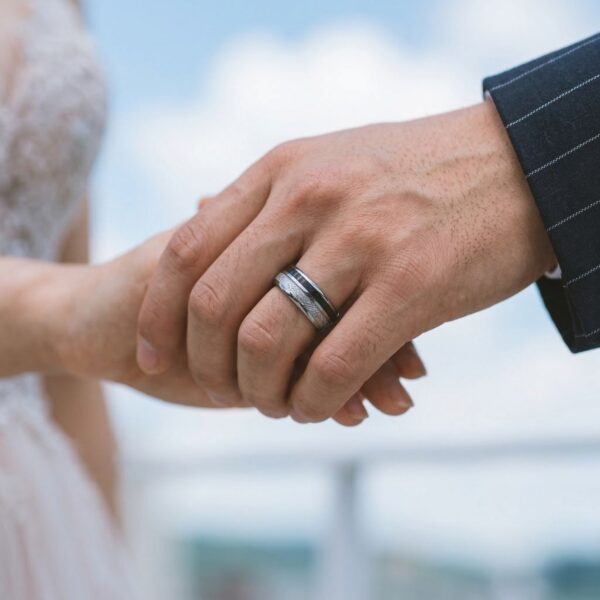 Newly married, the bride and groom reach for each others hands. The image shows the hands in a close up image. The man is wearing a silver tungsten ring which features two striking inlays. One of faux meteorite and the other of black carbon fibre.