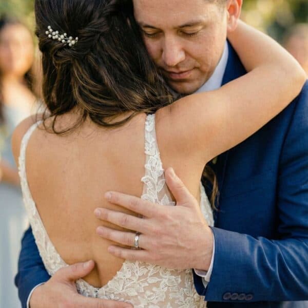 Holding each other, the newly wedded couple enjoy an embrace together. The bride has her back facing the camera, the groom faces forward. As he holds his new wife, he is wearing is silver tungsten thin style wedding ring.