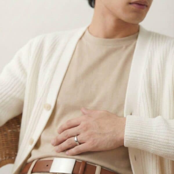 Sitting back in a very relaxed pose and dressed in light colours, the man has his hand across his stomach. He is wearing a thin style silver tungsten ring.