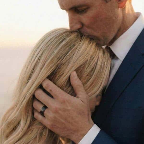 A newly wedded couple embrace, the man kissing his wife on the top of her head. On his hand he wears a black ceramic wedding ring.