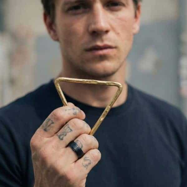 Holding a gold triangle, a man faces the camera. With tattoos on his hand, he wears a black ceramic ring.