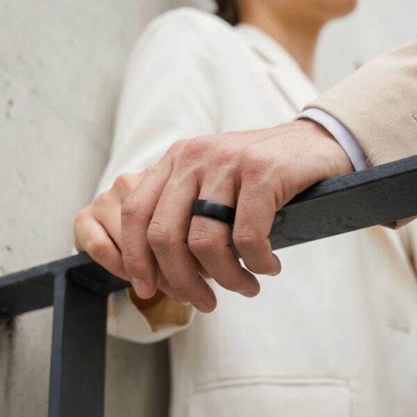 Holding onto the railings, a couple stand together, their hand on top of each others. The man is wearing a brushed black ring made with ceramic.