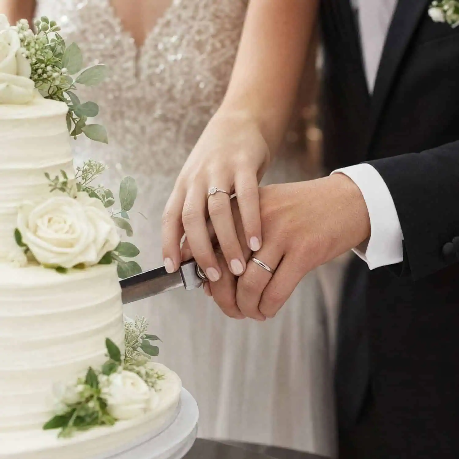 Bride and groom cutting their tiered wedding cake decorated with white flowers, with the groom wearing a thin silver tungsten wedding band and the bride wearing a diamond ring.