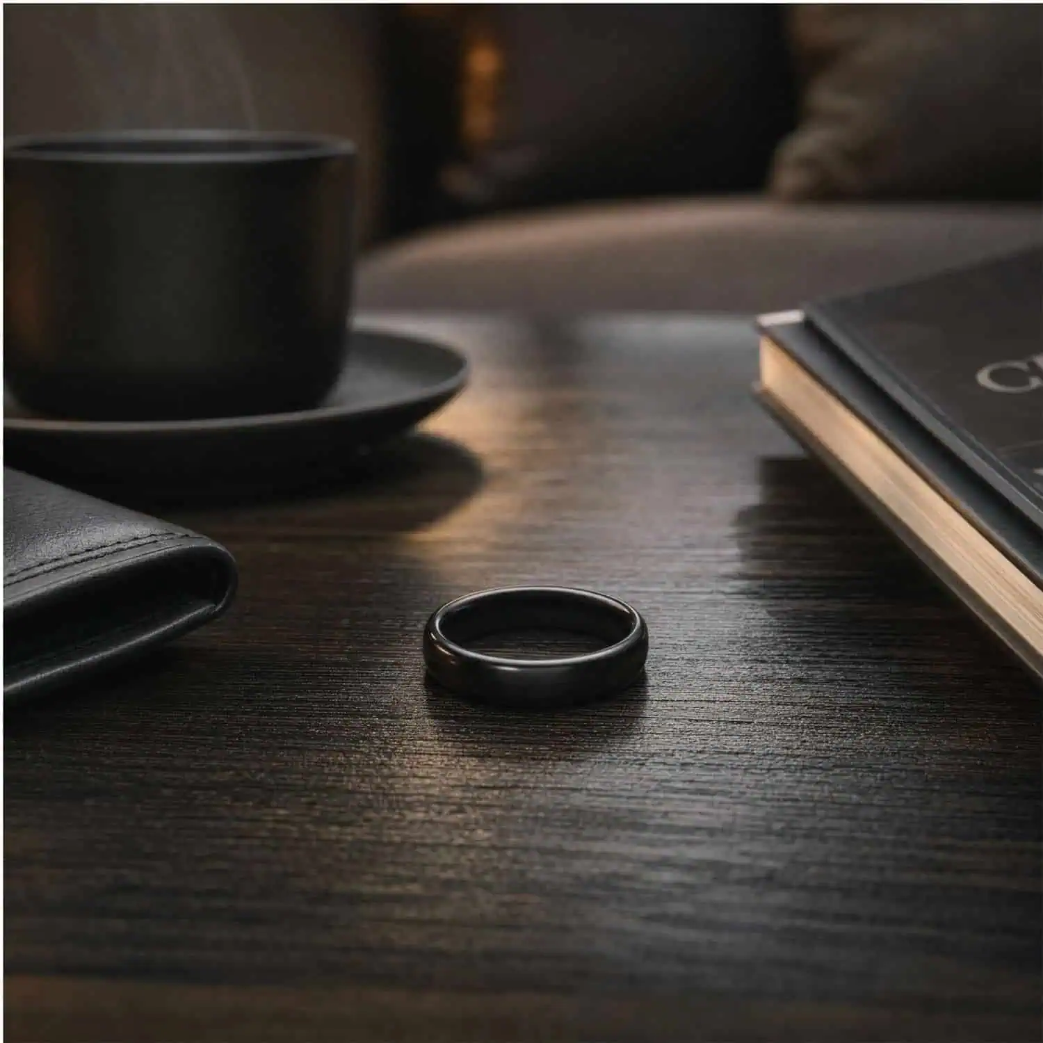 Men's polished black ceramic ring lying flat on a dark coffee table beside a black coffee cup and a small stack of books.