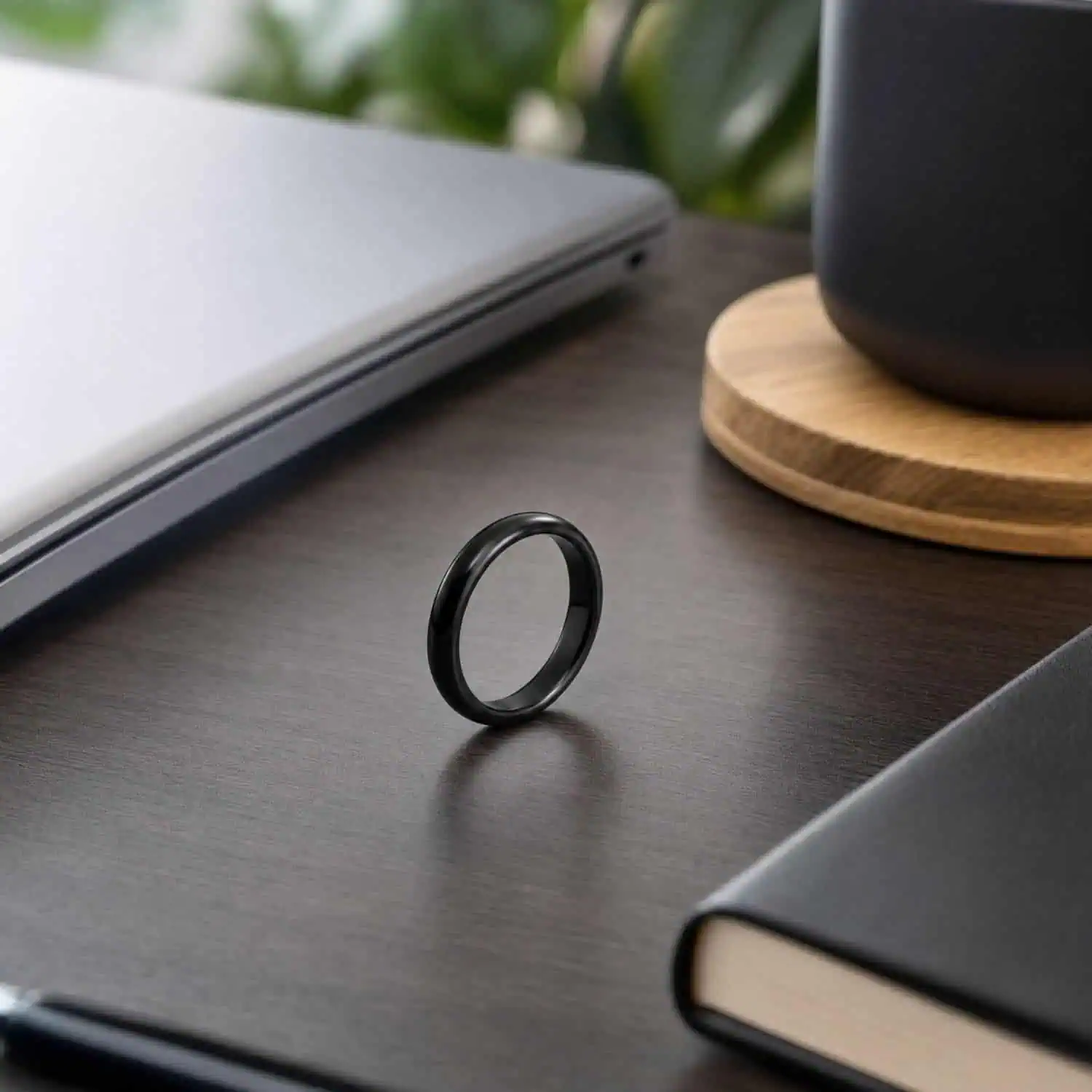 Polished black ceramic men's ring standing upright on a modern office desk beside a closed laptop, book and speaker.