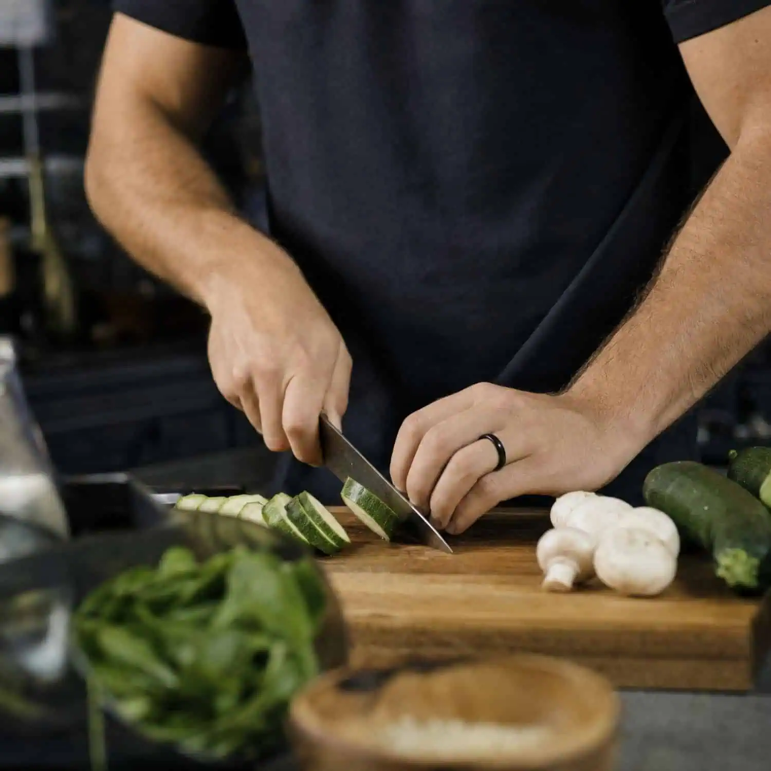 Man wearing a thin polished black ceramic ring while chopping courgettes and mushrooms on a wooden chopping board, dressed in a black T-shirt.