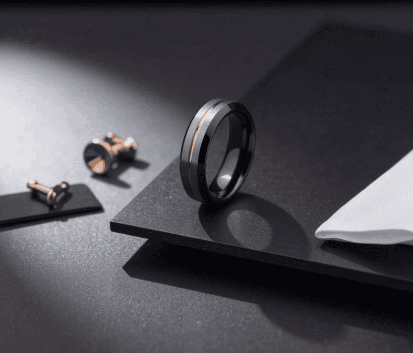 Stylish black tungsten ring with a subtle rose gold inlay, displayed on a modern dark surface alongside elegant accessories.