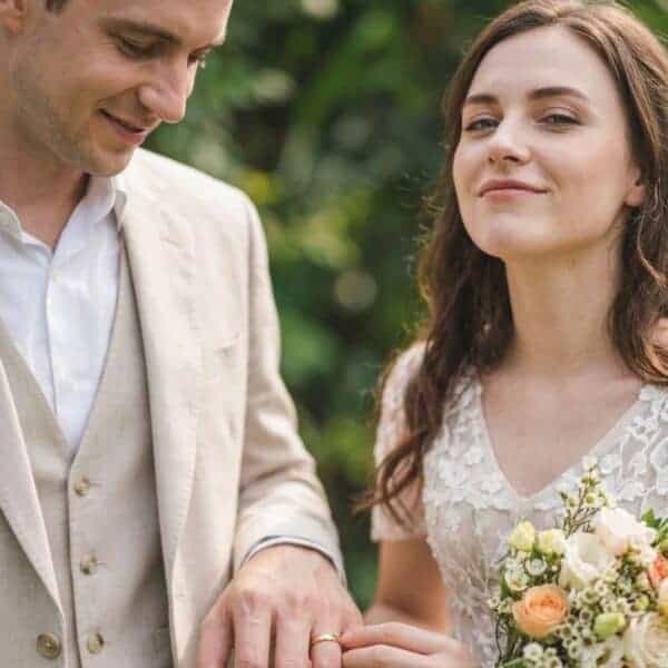 The newly married couple hold hands. The bride is wearing a white wedding gown and is holding white and light orange flowers. The man is dressed in a white shirt and a cream coloured suit. He is wearing a slim style gold tungsten wedding ring.
