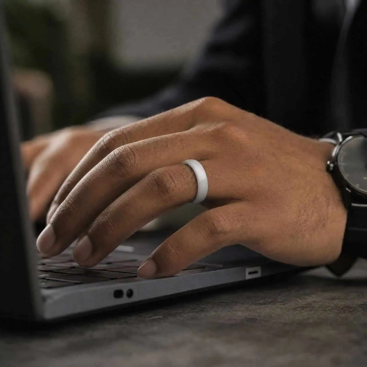 Close-up of a man working on his laptop while wearing a polished white fashion ring, with a dark shirt and wristwatch visible.