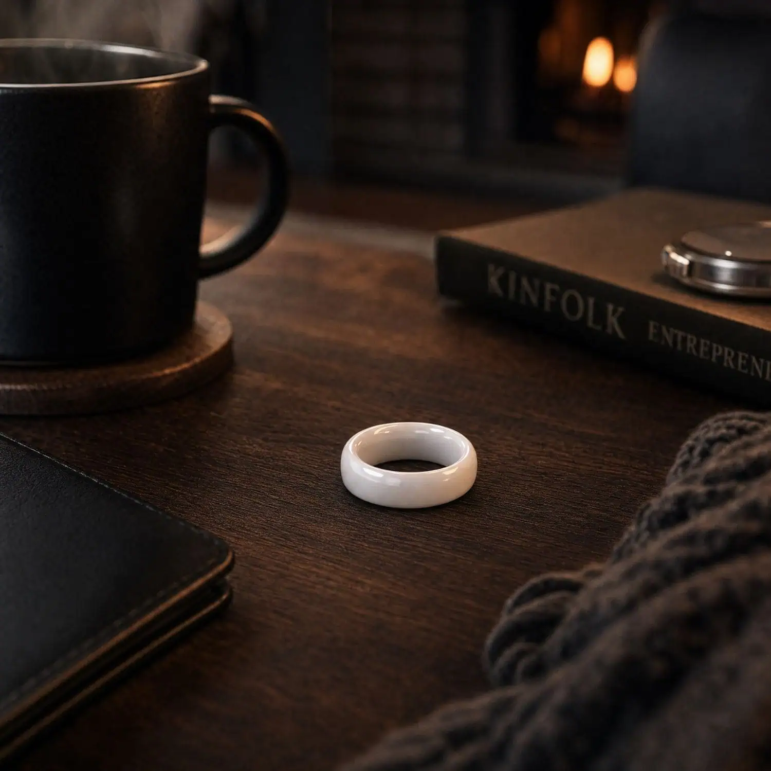 Polished white men's ring lying flat on a dark wooden coffee table beside a steaming cup of coffee, an open book and a leather-bound notebook.