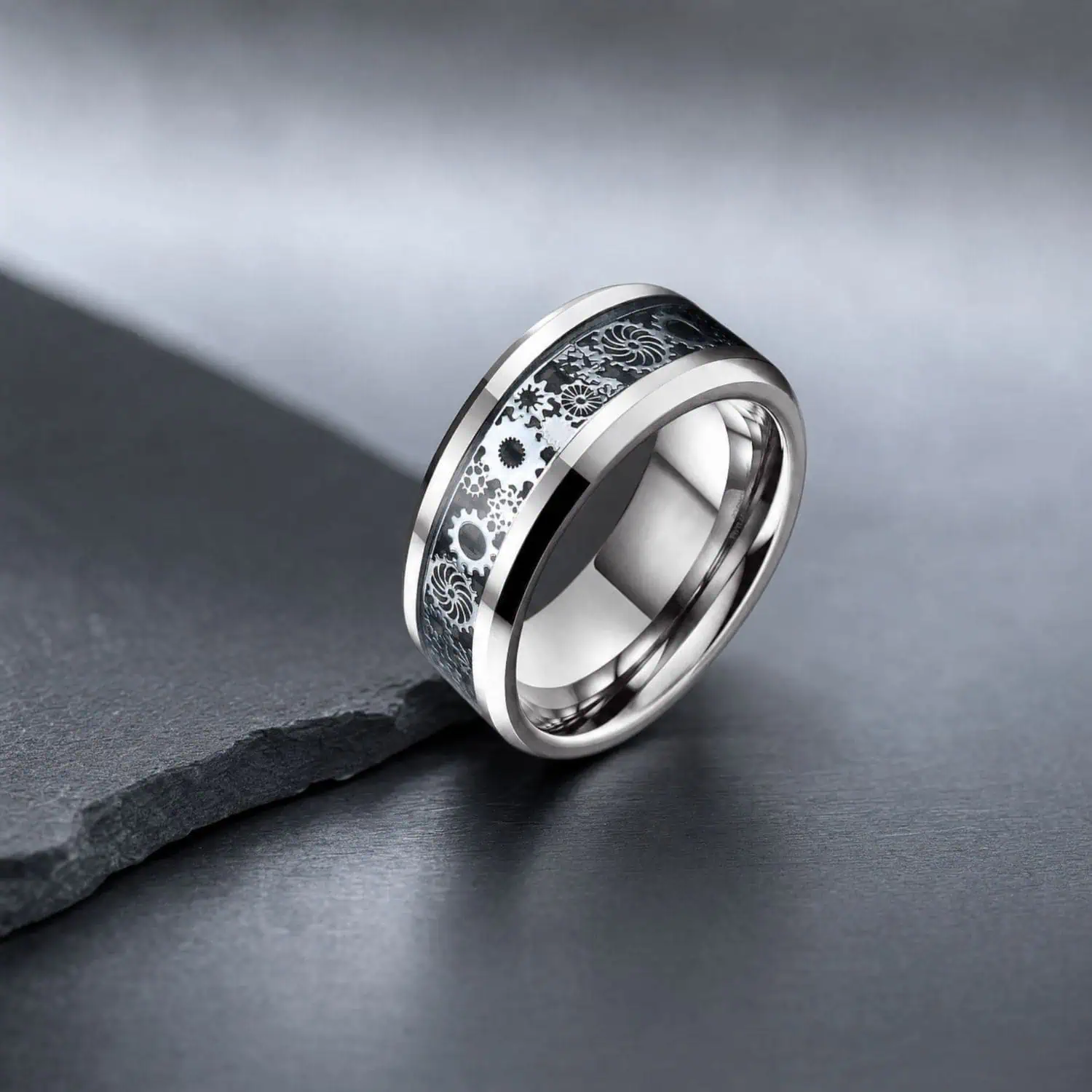 Black carbon fibre tungsten ring standing upright on charcoal slate, showing the polished silver band and intricate gear detailing beneath the glossy inlay.