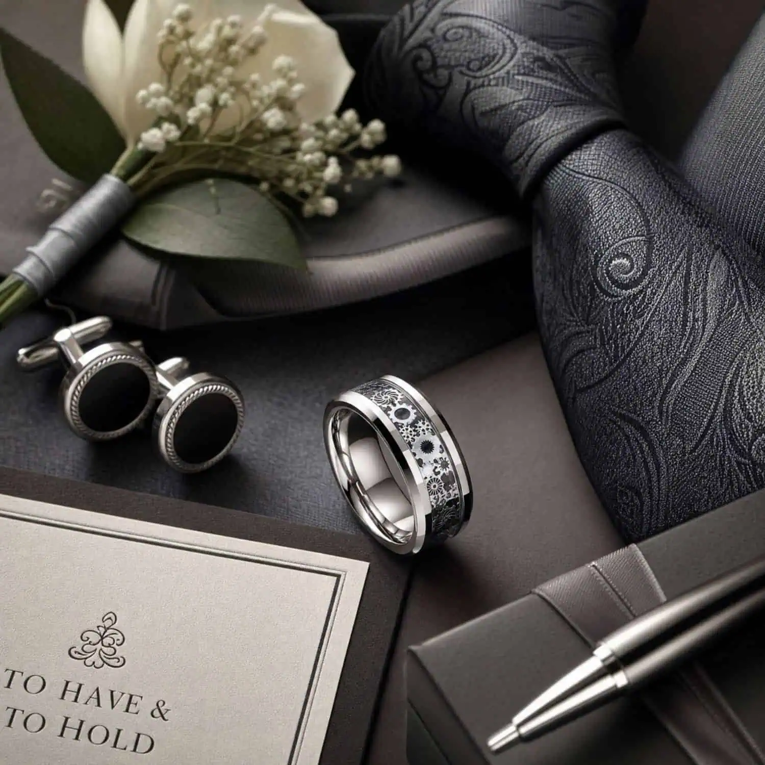 Carbon fibre tungsten wedding band displayed with the groom's vows, silver pen, tie, cufflinks and white flowers ready for the wedding ceremony.