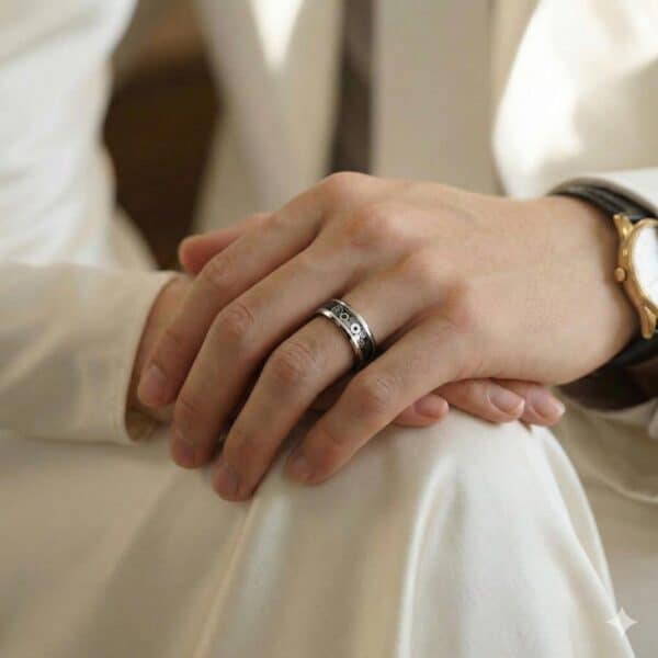 Dressed in white, a man sits with his hands resting on his knees. Wearing a gold watch, he has a ring on his hand. It is a silver and black tungsten ring with a carbon inlay that has a cogwheel design.