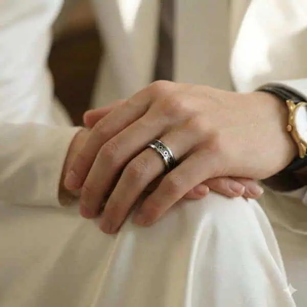 Dressed in white, a man sits with his hands resting on his knees. Wearing a gold watch, he has a ring on his hand. It is a silver and black tungsten ring with a carbon inlay that has a cogwheel design.