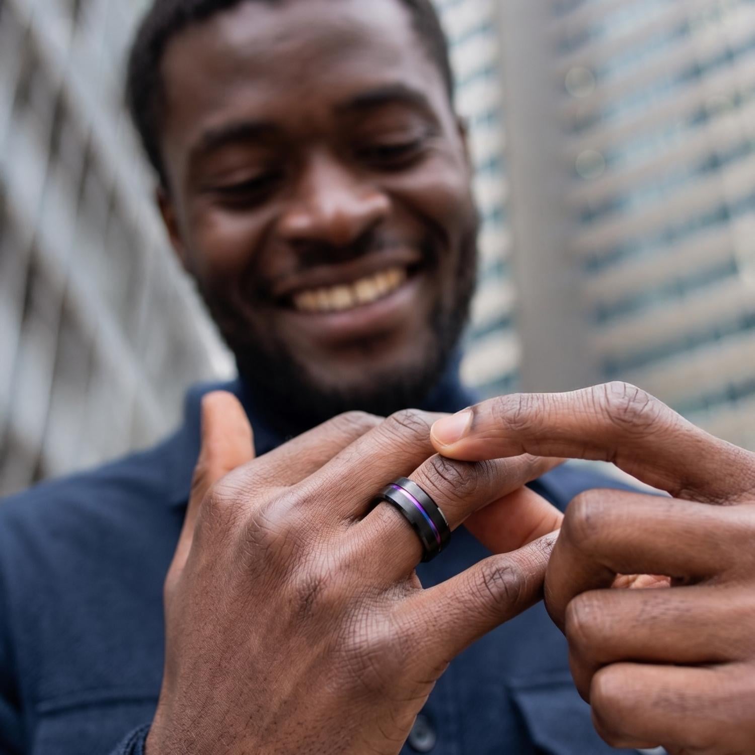 Man wearing the Unity men's black tungsten ring with an iridescent rainbow groove, smiling as he admires the ring with skyscrapers in the background.