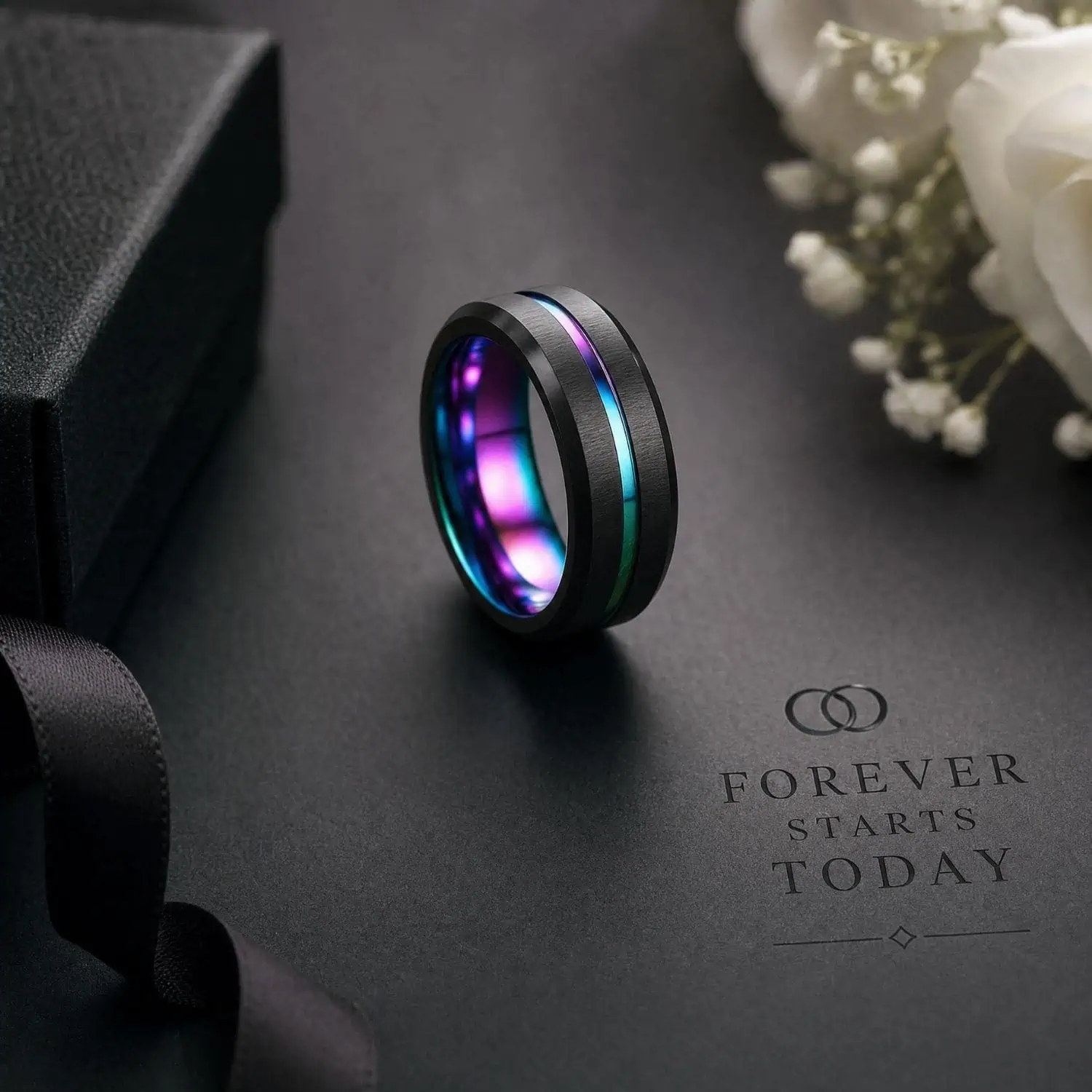 Black tungsten wedding ring with an iridescent rainbow groove displayed beside the words 'Forever Starts Today', white wedding flowers, a black ribbon and ring box.