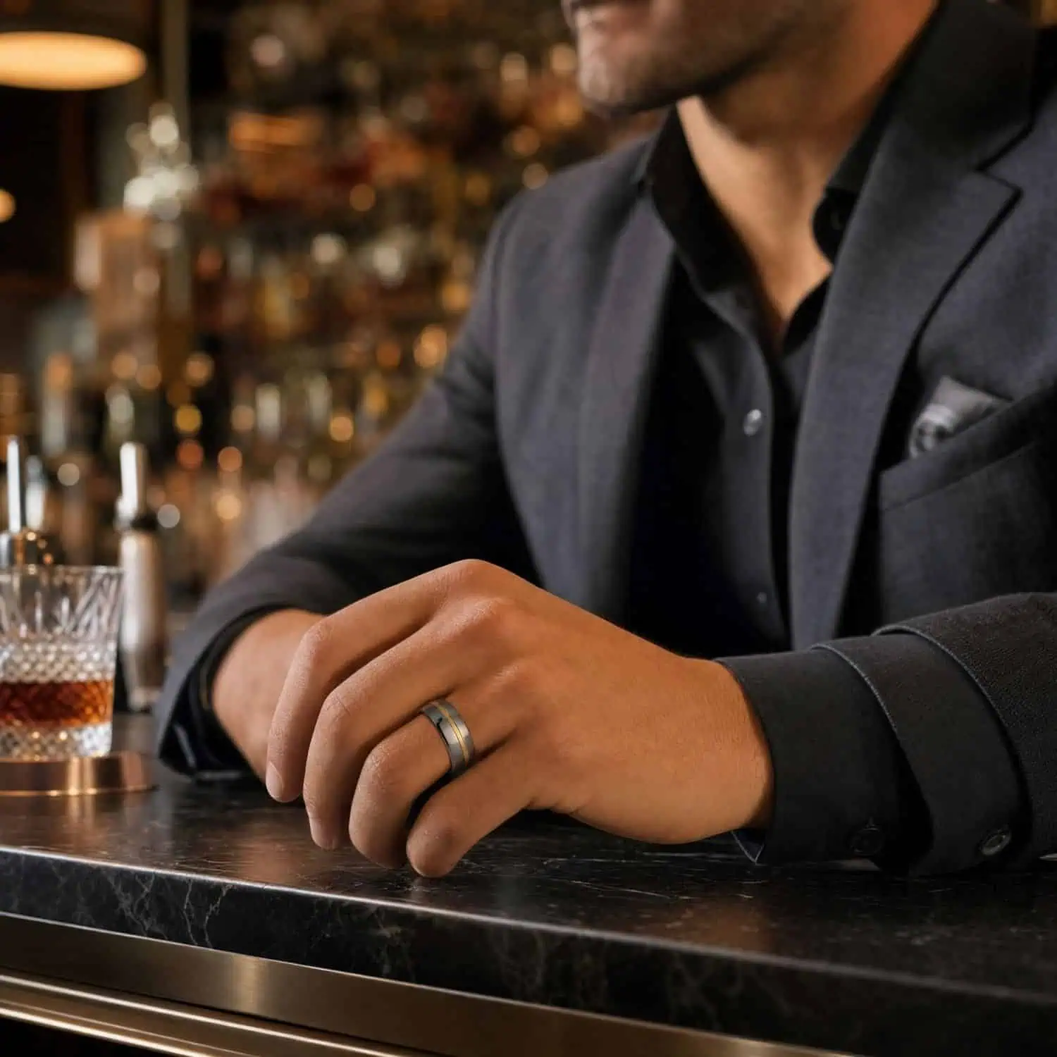 Man wearing a polished silver tungsten ring with a gold centre groove while resting his hands on a marble bar beside a glass of whiskey.