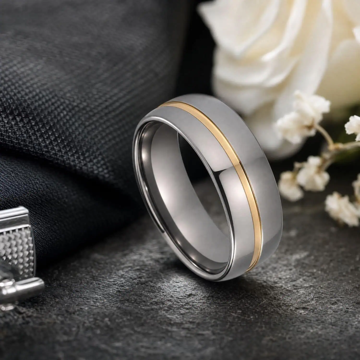 Two-tone tungsten wedding band in polished silver with a gold centre groove, displayed on grey slate beside white wedding flowers and cufflinks.