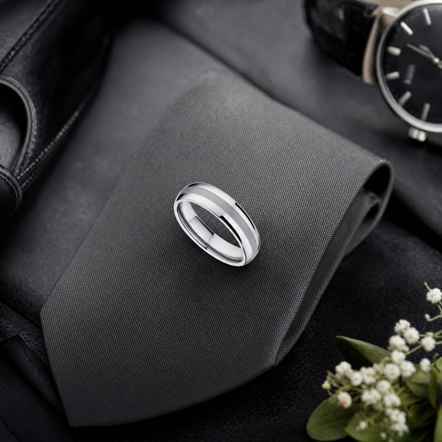 The Knight brushed silver tungsten wedding ring displayed on the groom's folded tie beside white wedding flowers and a black and silver watch.