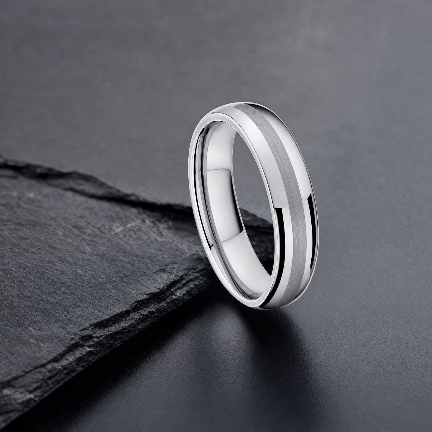 The Knight tungsten ring resting against dark slate on a minimalist grey background, highlighting the brushed centre and polished silver sides.