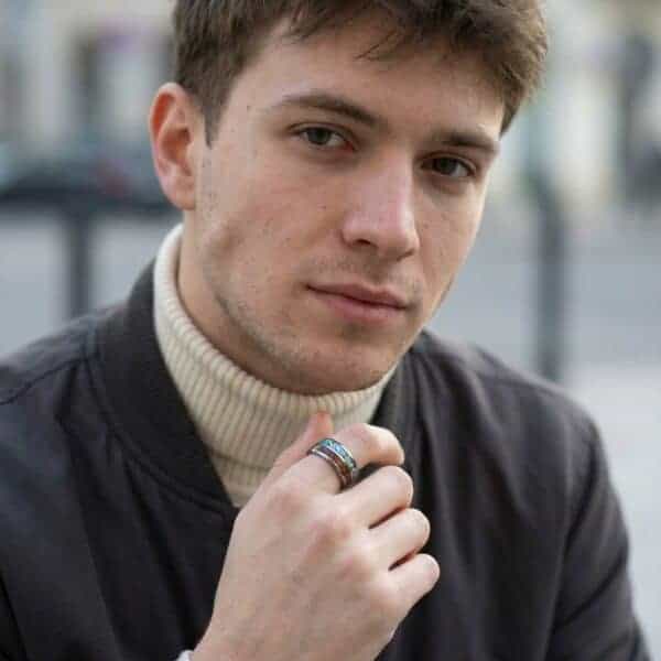 A man looks into the camera. Wearing a blazer and polo neck, he has his hand in front of his chest. On his hand, he wears a silver tungsten ring that features inlays of abalone shell and natural wood.