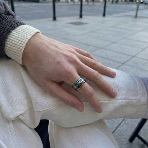 Resting on a chair, a man sits with his leg outstretched. With his hand on his knee, he has a tungsten ring on this hand. The ring features inlays of natural wood and abalone shell.