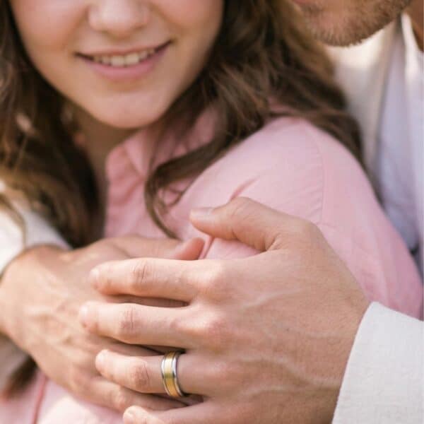 Enjoying a warm embrace, the young woman is smiling. The mans hands are close to the camera. He is wearing a gold and silver tungsten ring.