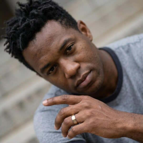 A close up of a mans face who is looking into the camera. He holds his hand in front of him, up towards his chin. On his wedding ring finger, he is wearing a gold and silver tungsten ring.