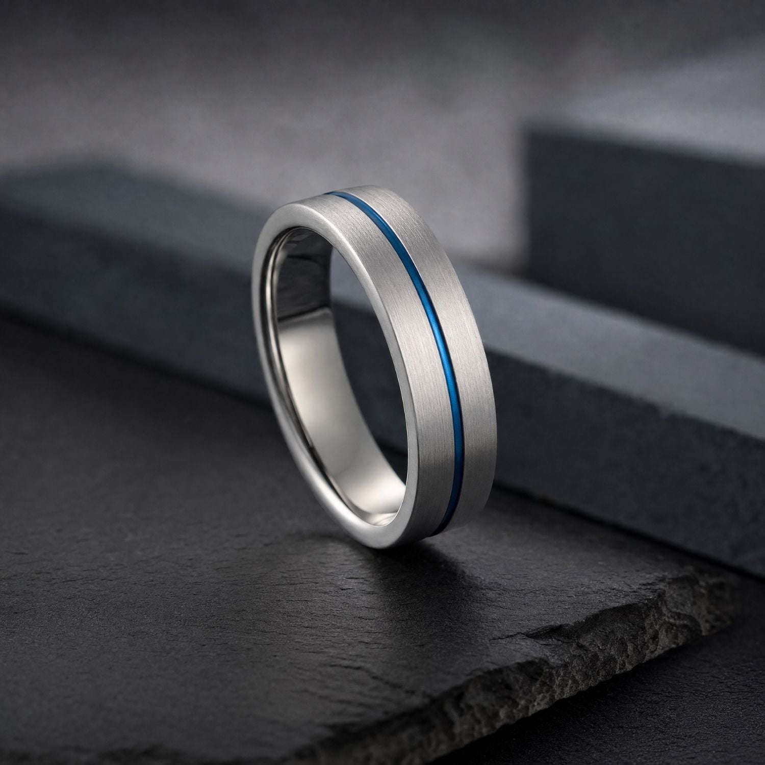 Negotiator brushed silver tungsten ring standing upright on dark grey slate against a minimalist background, highlighting its polished blue groove.