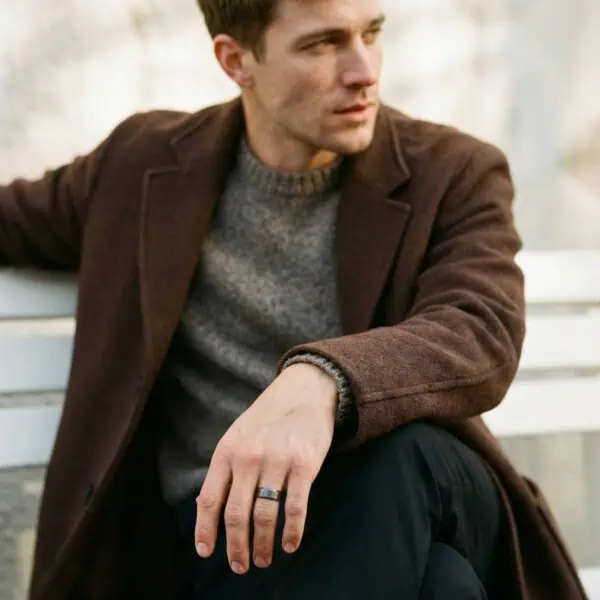 Sitting on a bench, the man sits in a relaxed pose. Wearing a casual jumper, and a brown blazer, he wears a ring on this finger. The ring is a gunmetal grey band that has a brushed centre and polished bevelled edges.