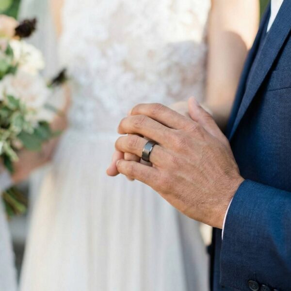 The couple join hands at the wedding ceremony. On the mans hand is a gunmetal grey tungsten ring - it has a brushed centre and polished bevelled edges.