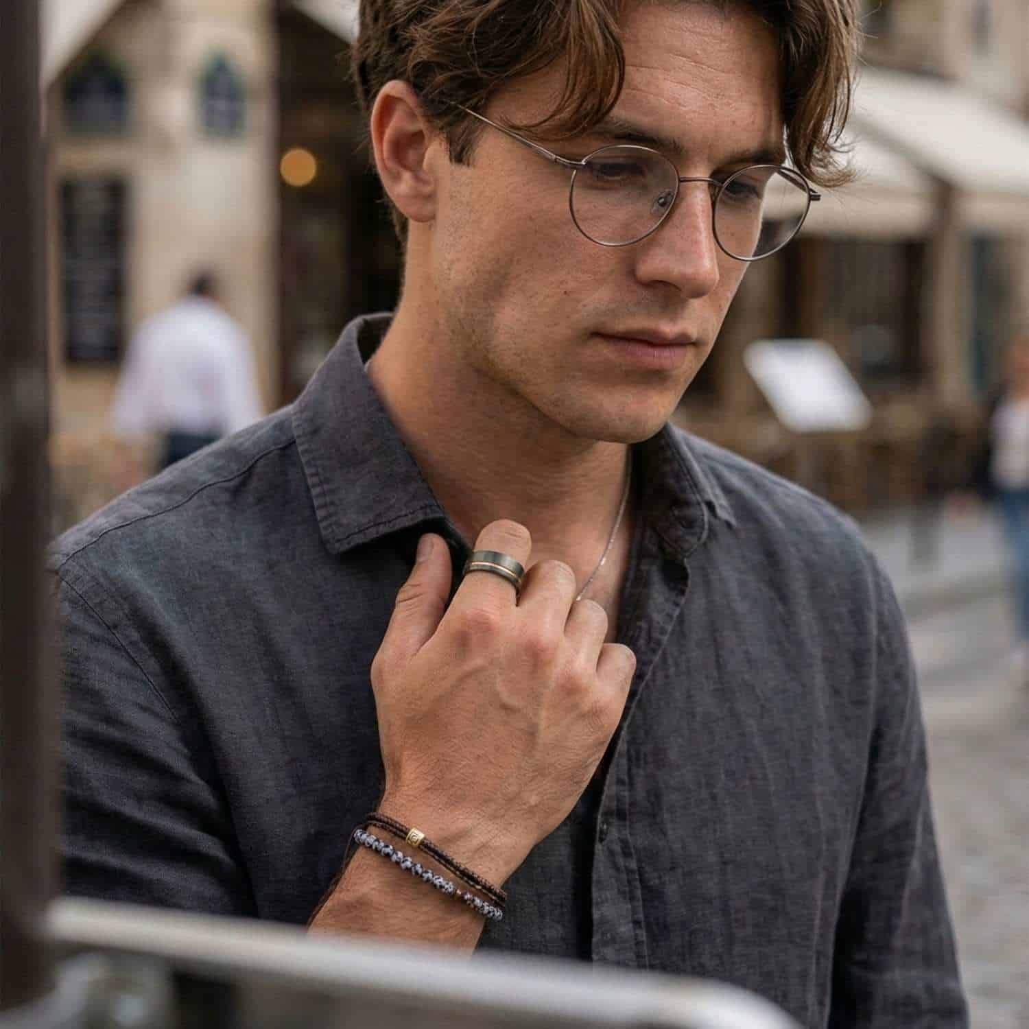 Out in the city, a man wearing a shirt brings his hand up to his chest. He is wearing two bracelets, and a tungsten ring. The ring is gunmetal grey with rose gold.