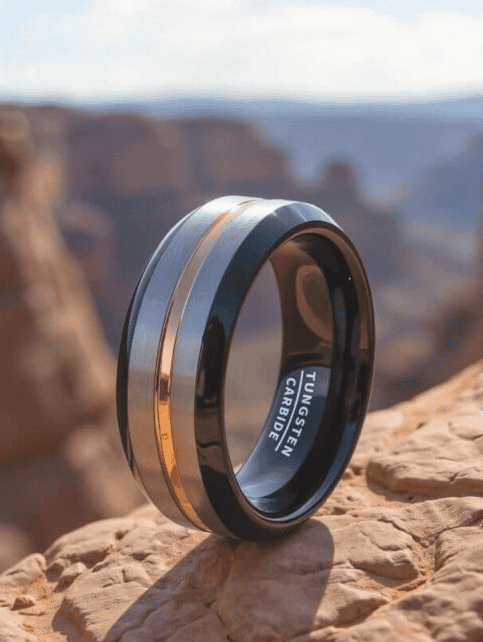 A stylish tungsten carbide ring with a sleek black finish and a rose gold inlay, placed on a rocky surface with a scenic background.
