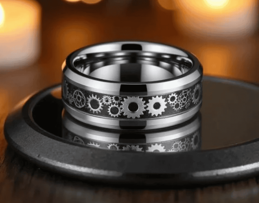 A polished tungsten ring featuring a detailed inlay of interlocking metallic gears, symbolizing mechanical precision and strength, displayed on a reflective black surface with warm candlelight in the background.