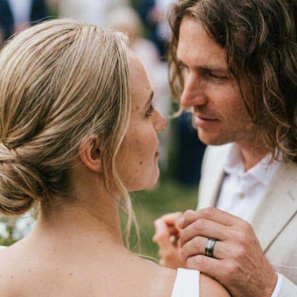 The bride and groom share a private moment together. Holding the brides and, the man is wearing his new wedding ring. It is a gunmetal grey tungsten ring and features an off centre groove or rose gold.