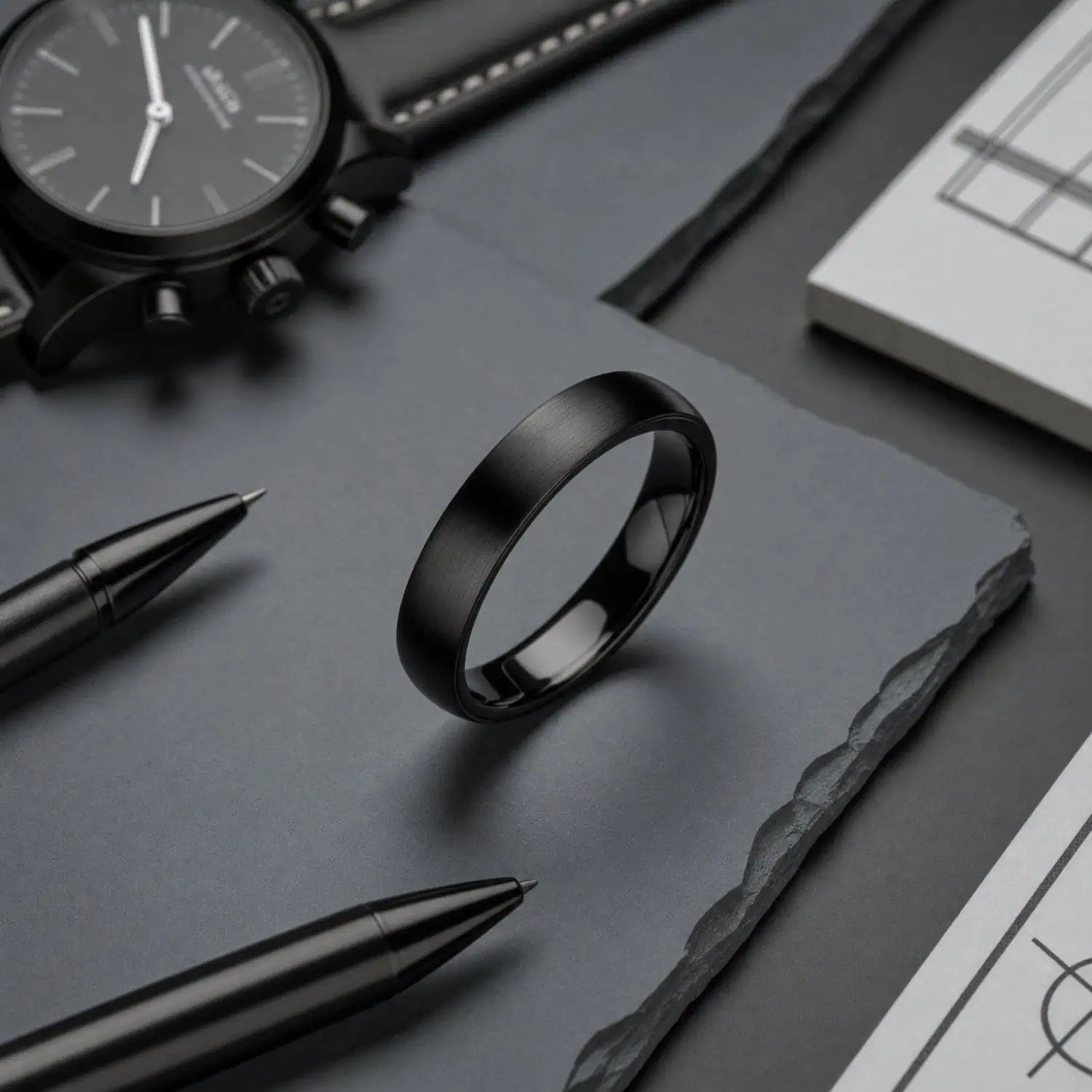 Raven men's black titanium ring standing upright on grey slate beside a watch and pens, highlighting its slim brushed black finish.