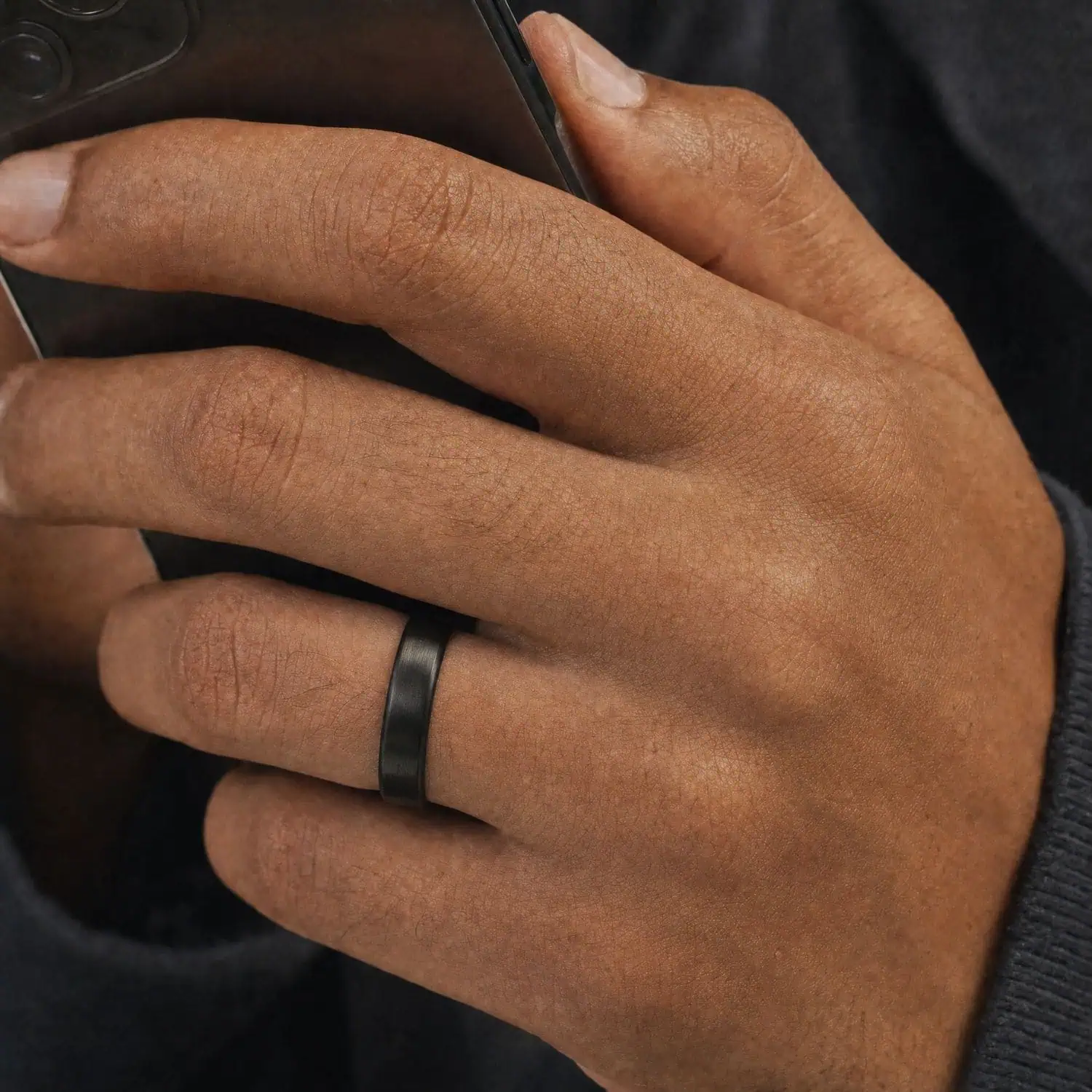Close-up of a man's hand holding his phone while wearing the Raven slim 4mm black titanium ring with a brushed matte finish.