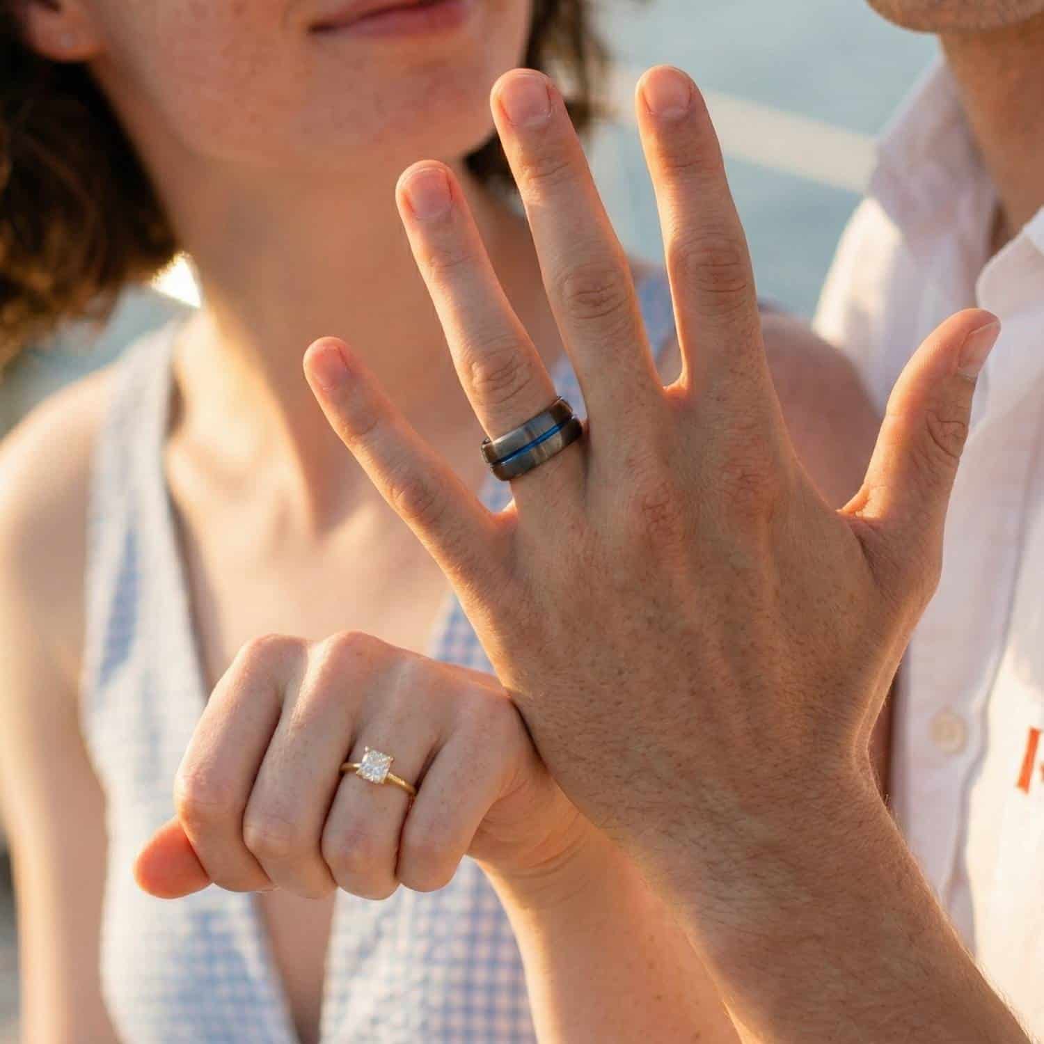 Holding up their hands to show their rings, the couple are delighted to show their rings. The woman has a diamond gold engagement band and the man has a gunmetal grey tungsten ring. It has a strip of blue running through the ring.