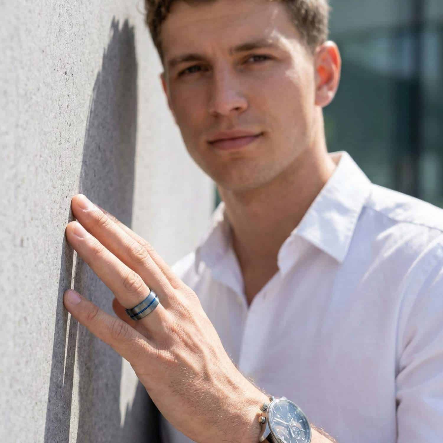 Leaning against a wall, a man wears a smart white shirt. On his hand he has a gunmetal grey tungsten ring that has a vivid strip of blue running just off centre of the ring.