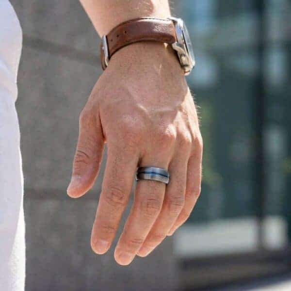 A close up image of a mans hand. One the middle finger, the man wears a ring. It is a gunmetal grey tungsten ring that has a groove of blue running through its centre.