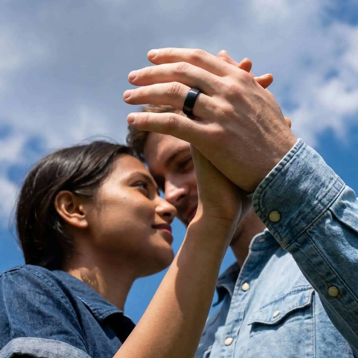 Happy couple holding hands beneath a blue but cloudy sky, with the man wearing the Valley black tungsten men's wedding band featuring a hidden blue interior.