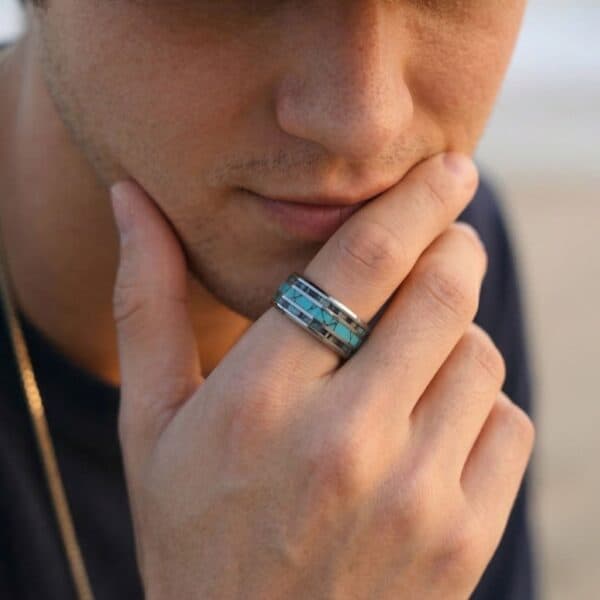 A close up shot of a young man who is holding his hand up to his chin and slightly covering his mouth. He is wearing a bold silver tungsten ring that is characterised by its turquoise inlay at the centre, and abalone shell inlays on either side.