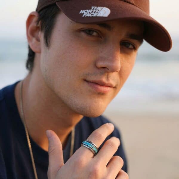 A young man smiles at the camera with a beach scene behind him. He wears a baseball cap and is holding his hand up in front of his chest. He is wearing a silver tungsten ring that features inlays of abalone shell and a centre inlay of turquoise.