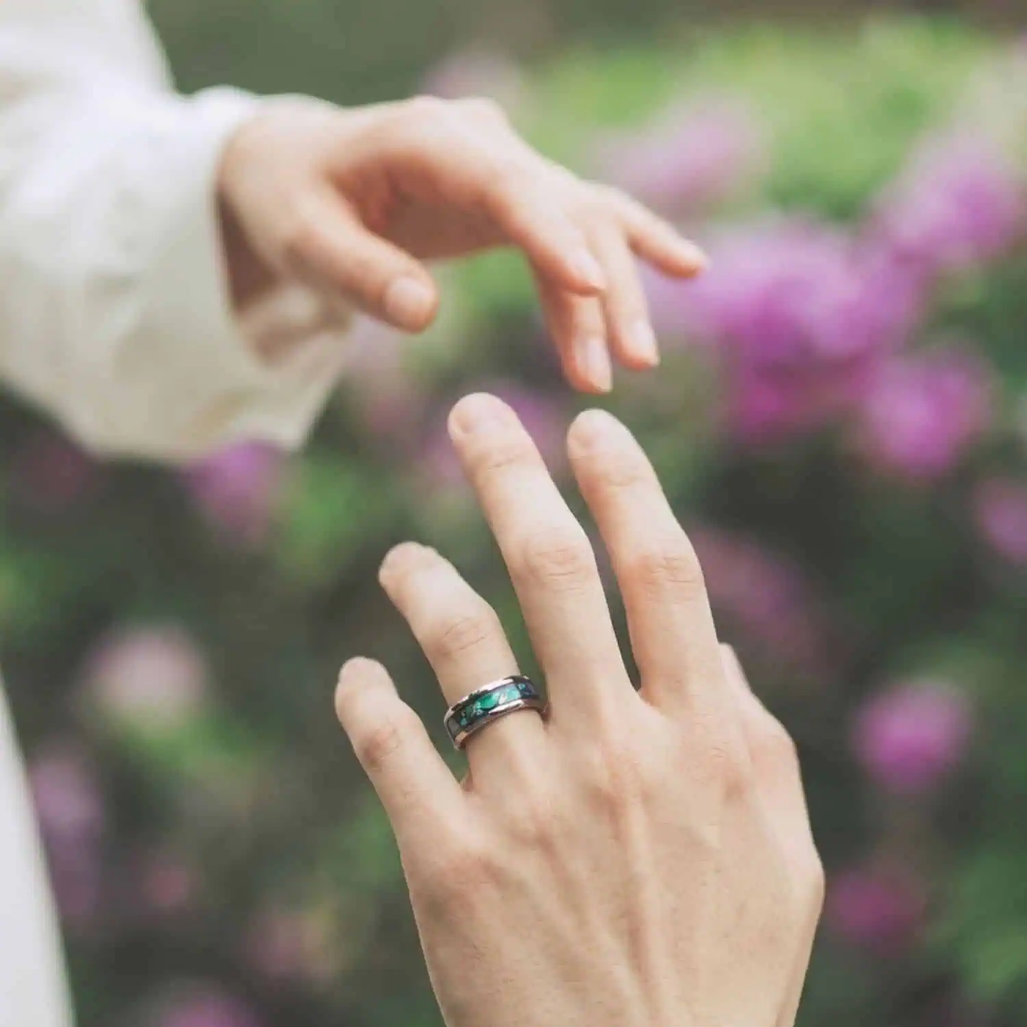 Bride and groom reaching out to hold hands in a garden wedding while the groom wears the Reef silver tungsten wedding band with an abalone shell inlay.