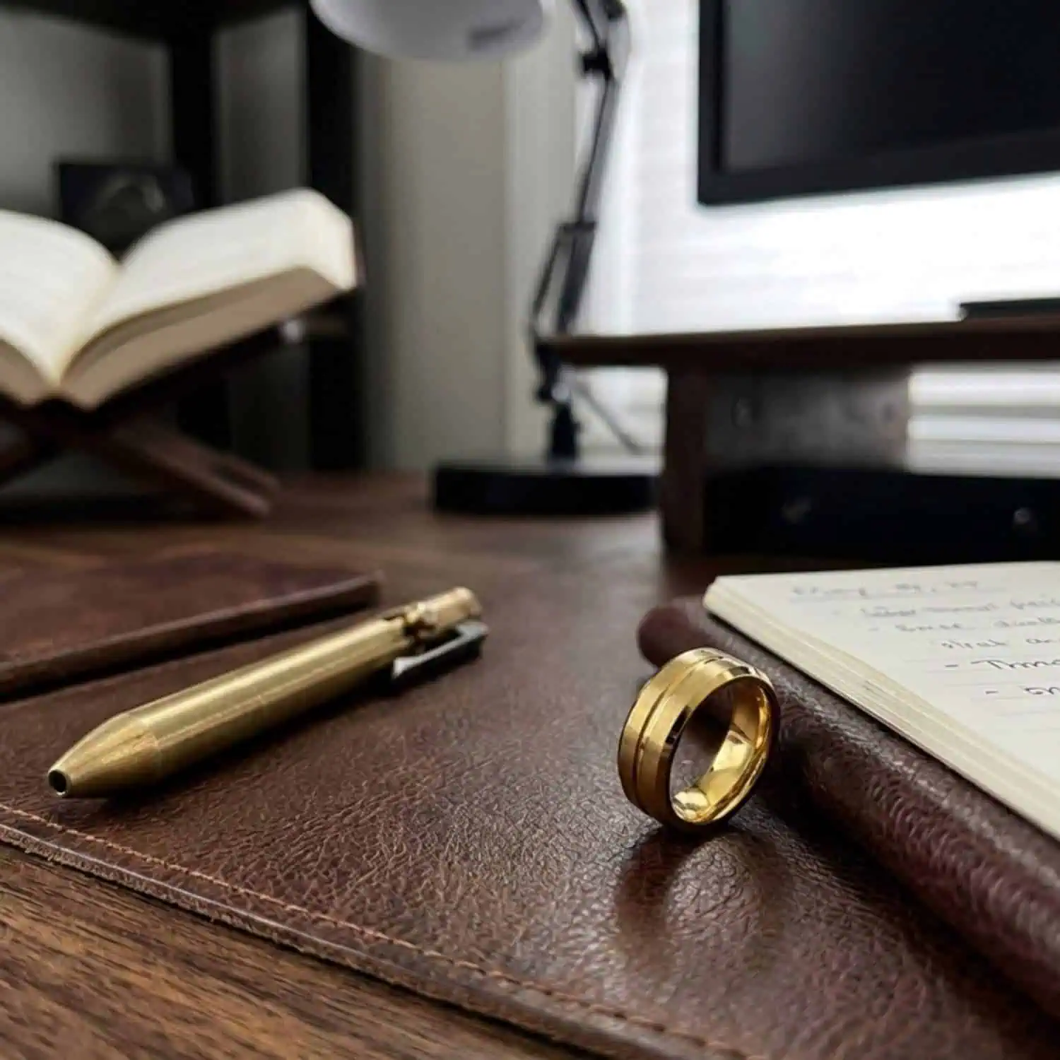 Valour gold tungsten ring displayed against a brown leather journal on a wooden desk, with a desk lamp and computer softly blurred in the background.