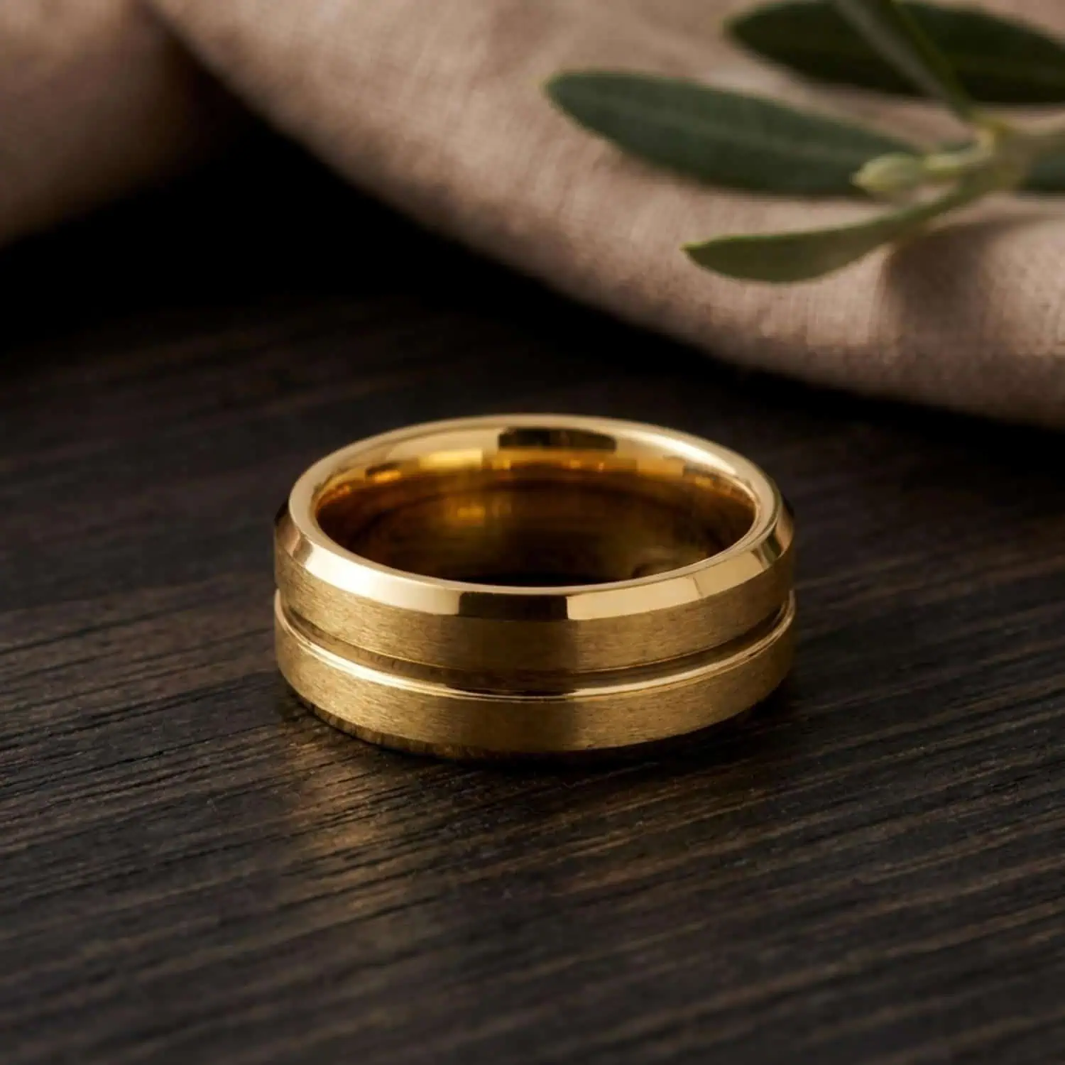 Valour gold tungsten ring standing upright on a wooden table with green leaves in the background, showcasing the brushed gold centre, polished centre groove, and polished bevelled edges.
