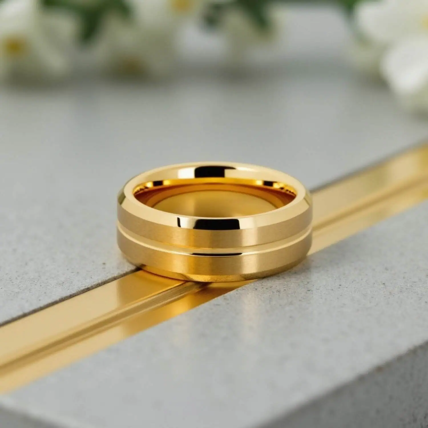 Men's gold tungsten wedding band with brushed raised sections, polished centre groove, and bevelled edges displayed with white wedding flowers in the background.
