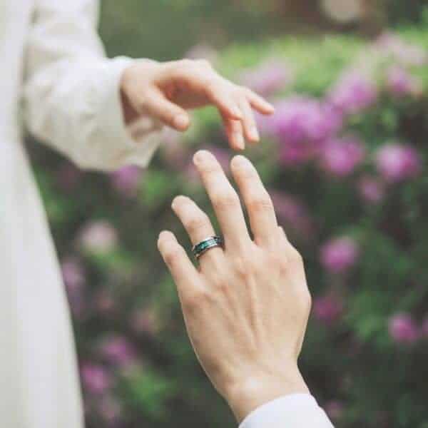 With blooming flowers in the background, a couple go to join hands. It looks like it is their wedding day as the woman is in a white wedding gown. The man has a ring on his wedding finger. It is a silver tungsten ring featuring abalone shell. With shimmering bevelled edges.